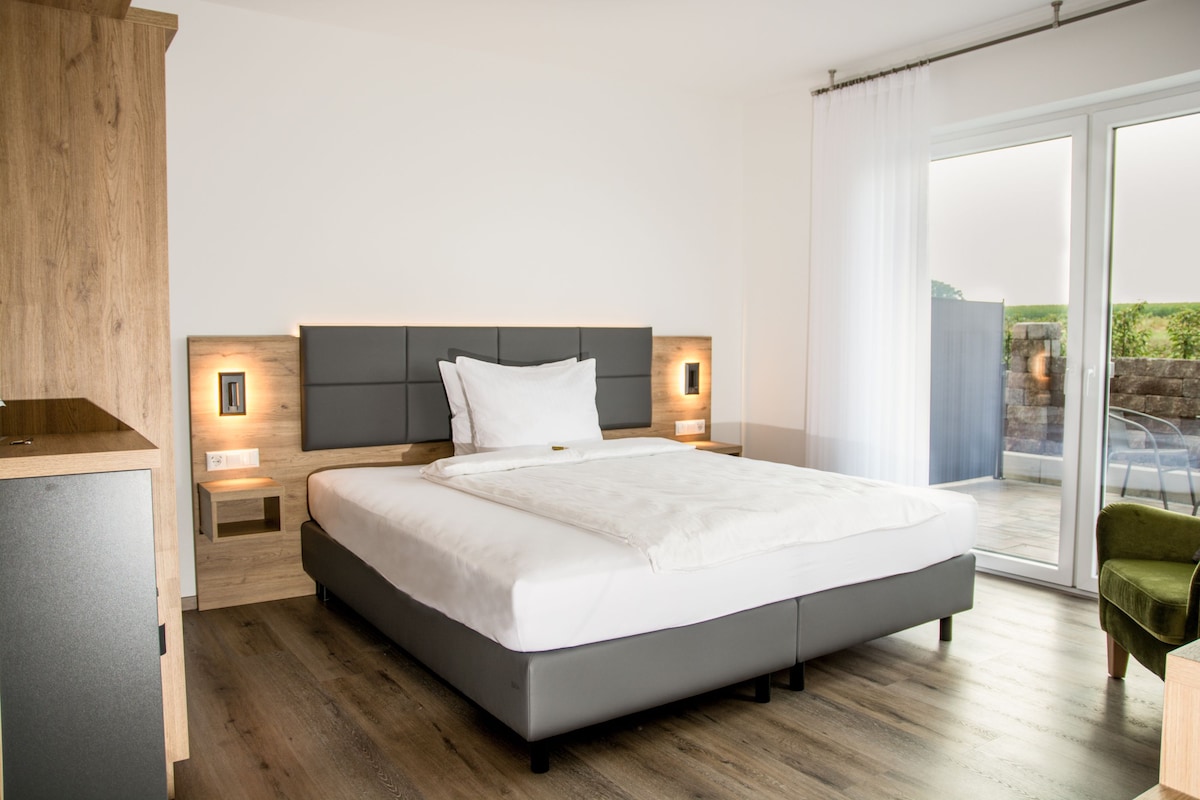 Une chambre moderne avec un grand lit, une tête de lit en bois, des tables de chevet avec des lampes et une vue sur l’extérieur grâce à une porte-fenêtre coulissante.