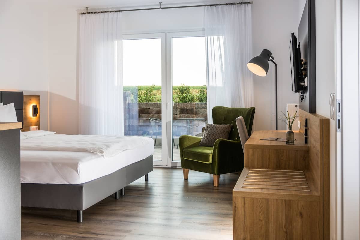Un dormitorio moderno con cama, un sillón verde, un escritorio de madera, televisor y un amplio ventanal con cortinas.