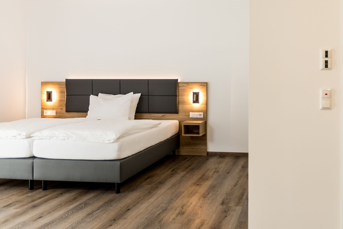 A modern bedroom with a grey bed, wooden headboard, bedside tables with built-in lighting, and a wooden floor.