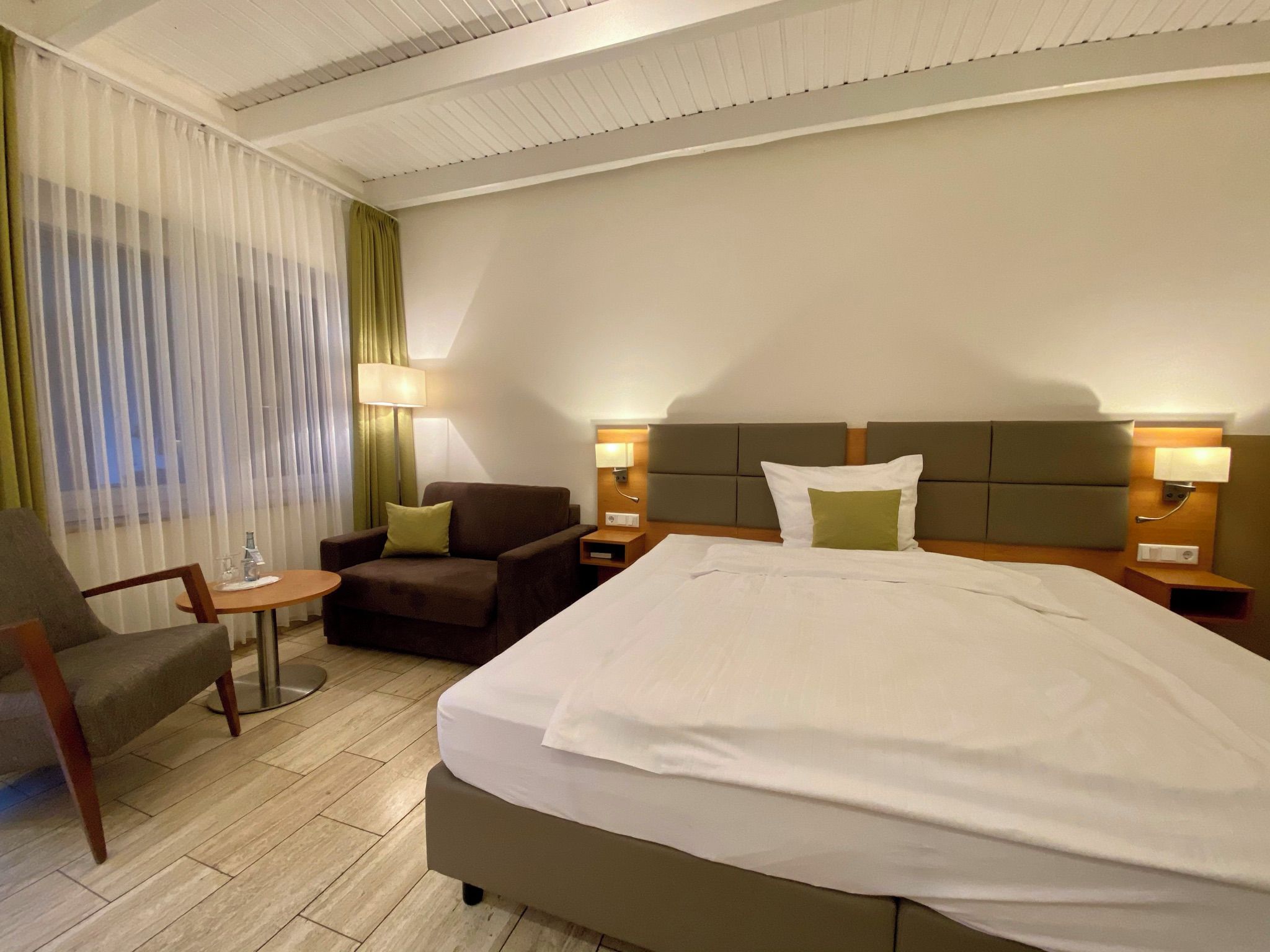 A hotel room with a large bed, a sofa, a chair, a small table, and a lamp.