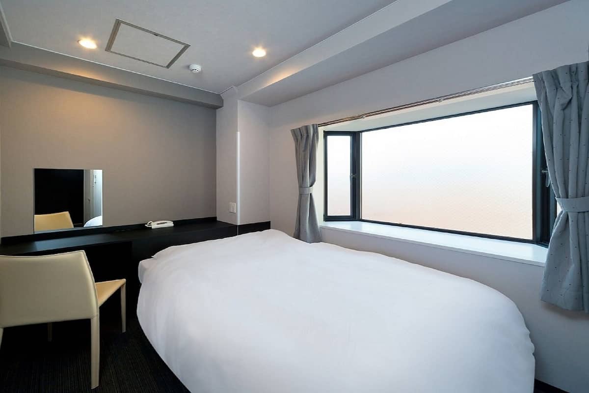 Une chambre d’hôtel avec un lit, un bureau, une chaise, un miroir et une fenêtre avec des rideaux.