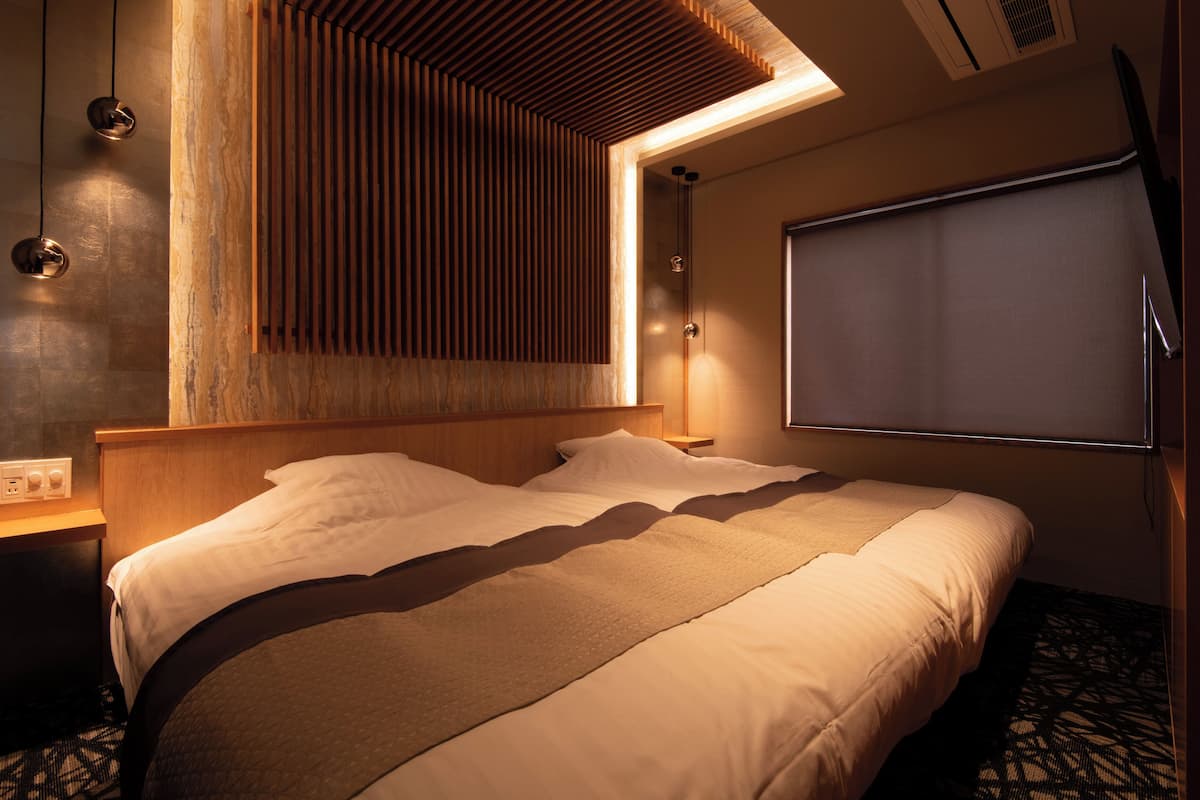 A hotel room with a bed, wooden headboard, bedside lamps, a wall-mounted TV, and a window with blinds.