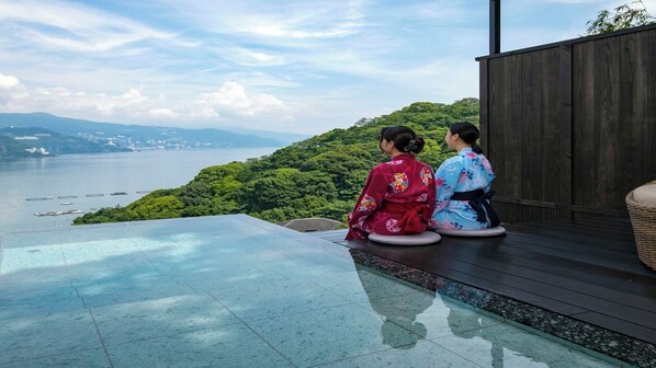 Two individuals in traditional attire sitting on a wooden deck overlooking a body of water and a mountainous landscape. - Jikan Wo Tabisuru Yado Umi No Hana (Atami)