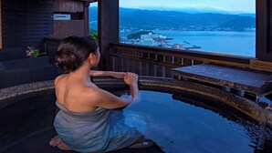 A person in a hot spring bath with a view of a body of water and buildings in the distance. - Jikan Wo Tabisuru Yado Umi No Hana (Atami)