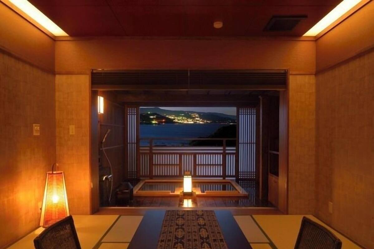 A room with a view of the sea, a wooden table, chairs, a lamp, and a traditional Japanese sliding door.