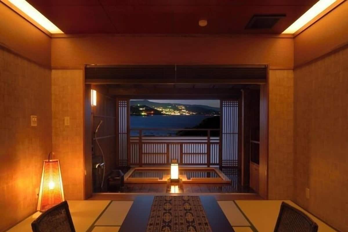 A room with a view of the sea, a wooden table, chairs, a lamp, and a traditional Japanese sliding door.