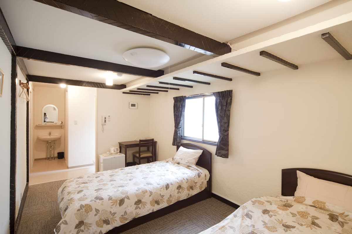 A bedroom with two single beds, a window with curtains, a desk, and a bathroom visible through an open door.
