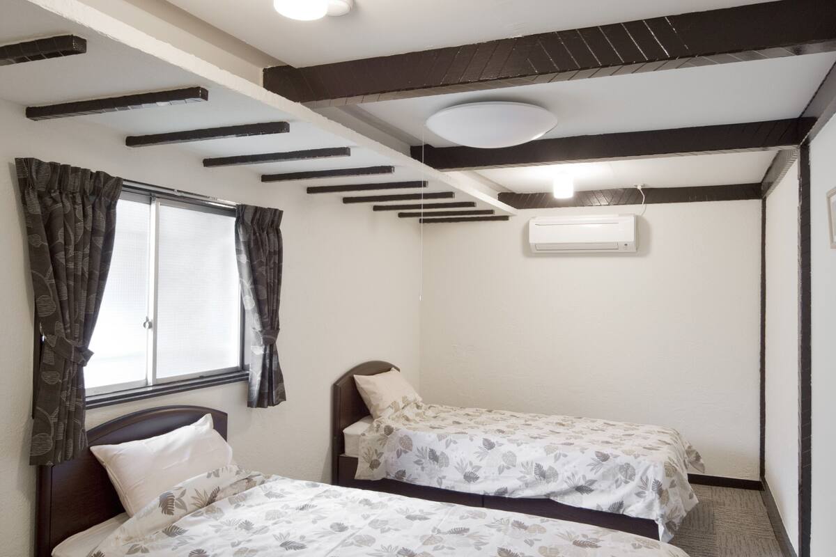 A room with two beds, a window with curtains, a ceiling light, and an air conditioning unit.