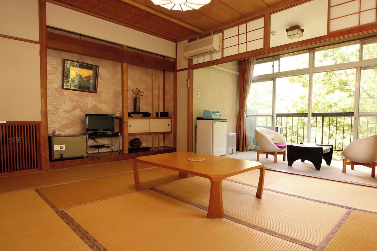 Ein traditioneller japanischer Raum mit Tatami-Boden, einem Fernseher, einem kleinen Kühlschrank und Blick auf Grün durch große Fenster.
