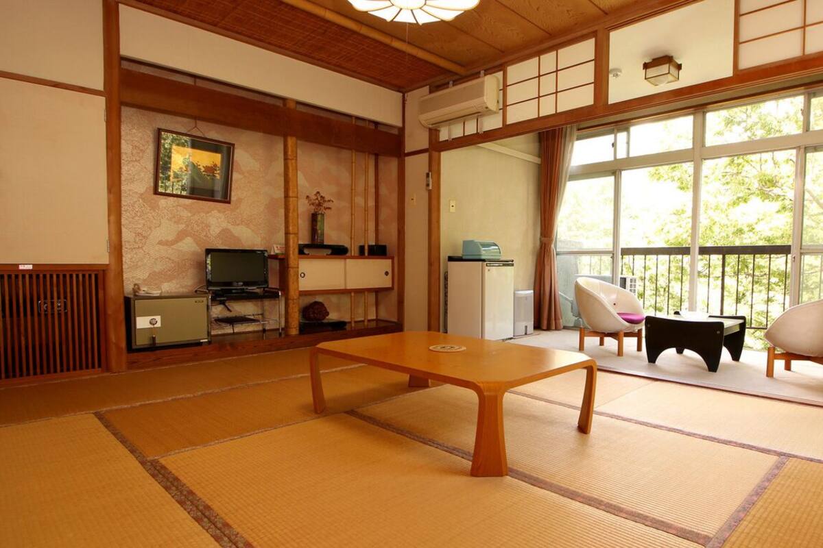Ein Raum mit Tatami-Matten, einem niedrigen Tisch, Stühlen, einem Fernseher und einem Fenster mit Blick auf Bäume.