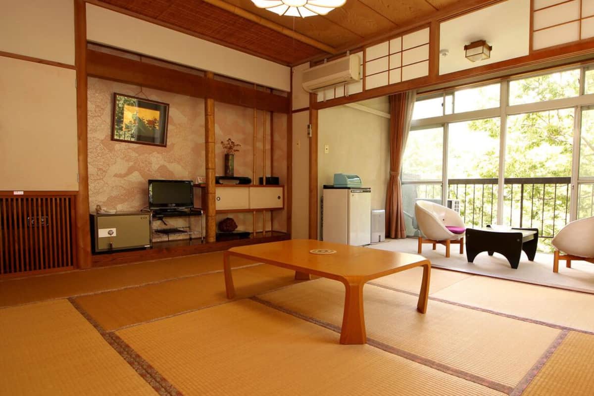 Una habitación con tatamis, una mesa baja, sillas, un televisor y una ventana con vistas a árboles.