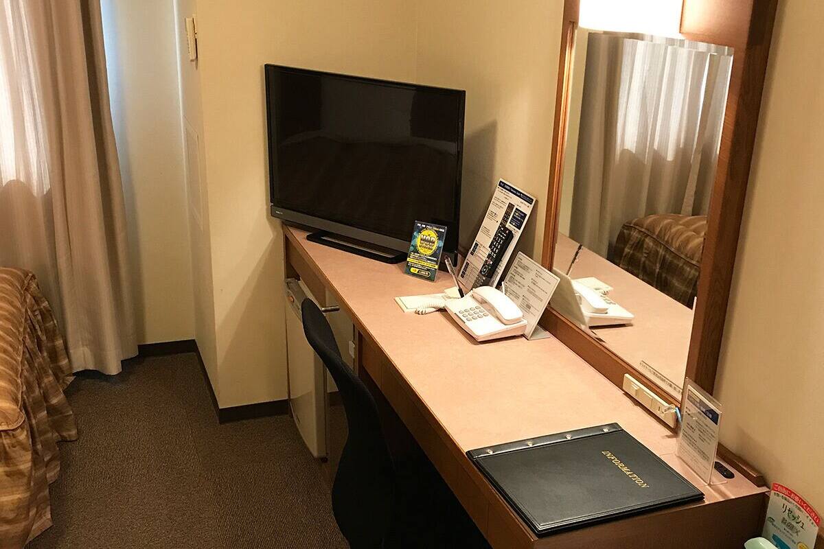 A hotel room with a desk, a television, a mirror, a telephone, and a small table with a book and a menu.