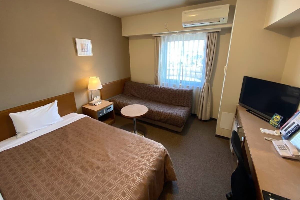 A hotel room with a bed, a sofa, a desk with a computer, a TV, and an air conditioning unit.