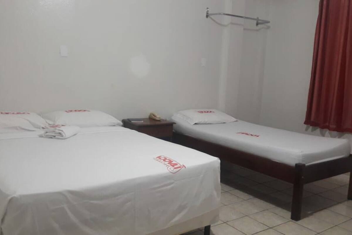 Una habitación pequeña y limpia con dos camas individuales, una mesita de noche, una cortina roja y un suelo de baldosas blancas.
