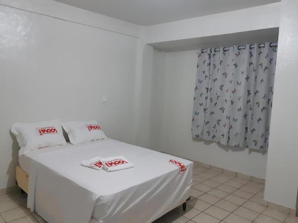 Uma cama de solteiro com colcha e almofadas brancas, uma janela com cortinas estampadas e piso de azulejos.