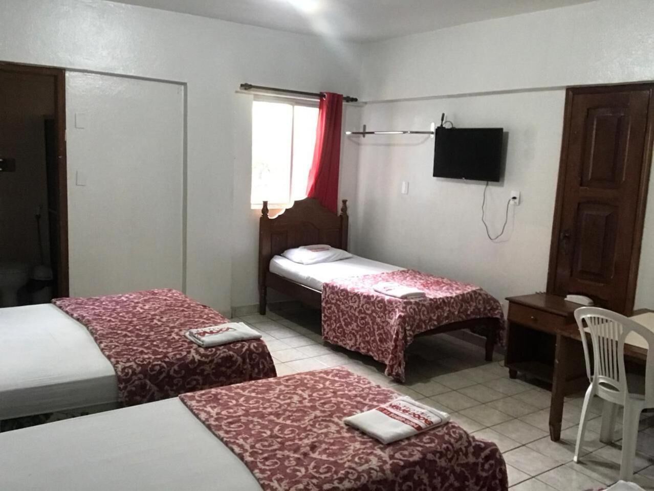 Ein Zimmer mit zwei Einzelbetten, einem Schreibtisch, einem Stuhl, einem Fernseher und einem Fenster mit roten Vorhängen.