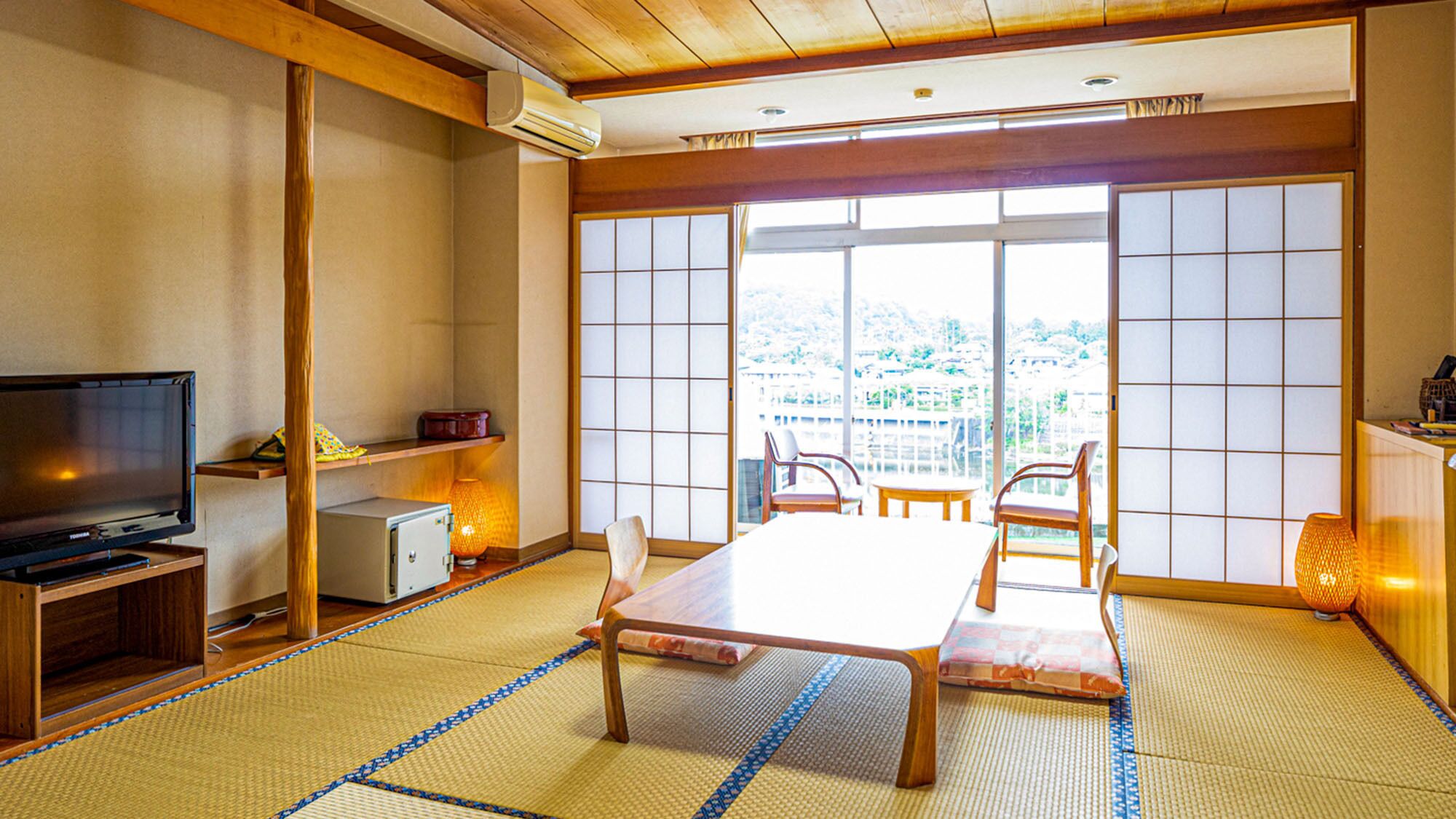 A traditional Japanese room with tatami flooring, a television, a small refrigerator, a table with chairs, and a view of a cityscape through large sliding doors.