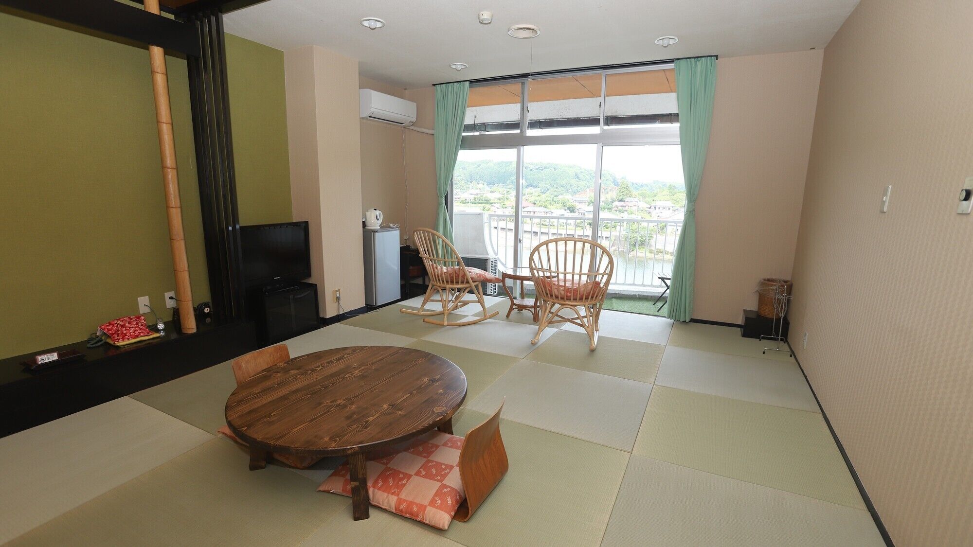 A room with tatami flooring, a round wooden table, two rocking chairs, a television, and a balcony with a view.