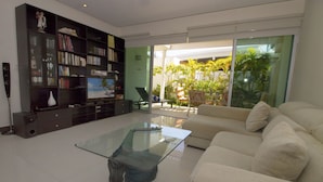 A living room with a glass coffee table, a beige sofa, a bookshelf, and a sliding glass door leading to a balcony with plants. - Downtown Apartment Oasis 12 (Playa del Carmen)