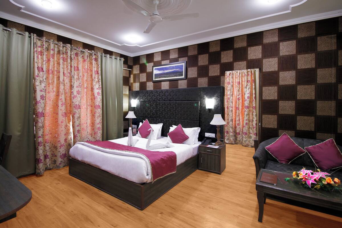 A well-lit hotel room with a large bed, a sofa, and a small table with a flower arrangement.