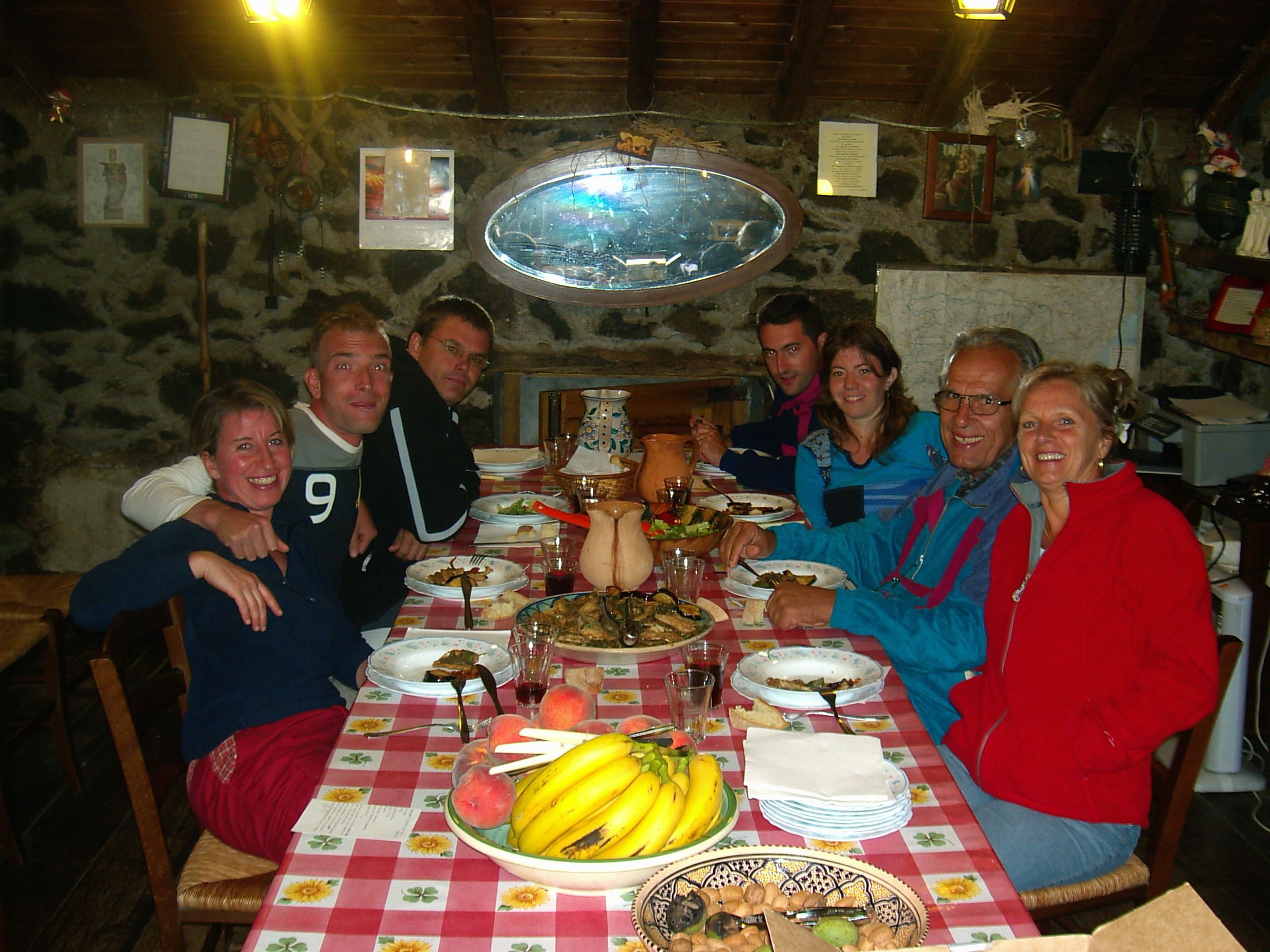 A group of people seated around a table with various dishes, including fruits and a pitcher, in a room with stone walls and a wooden ceiling.