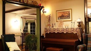 Una habitación acogedora con un piano, un sillón verde, un sillón escocés y una mesa con mantel de encaje.