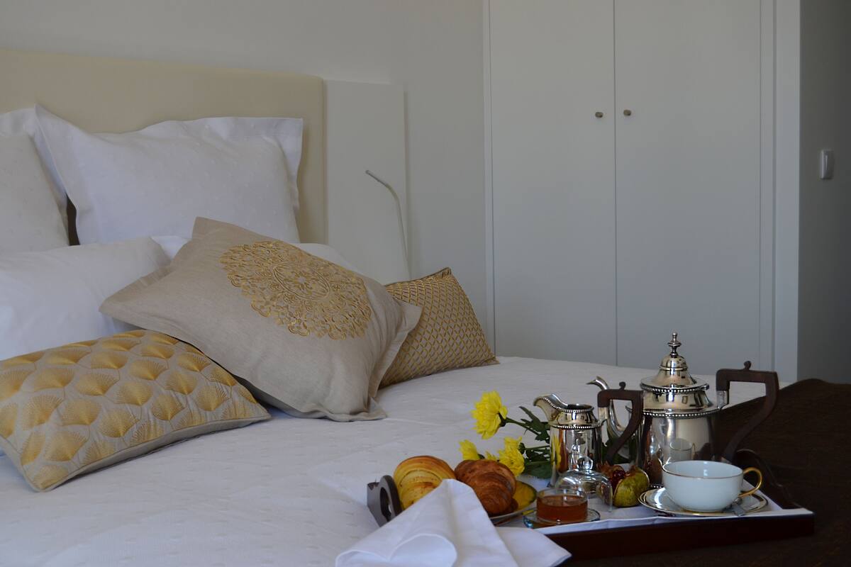 A neatly made bed with white and patterned pillows, a tray with croissants, a teapot, and a cup.