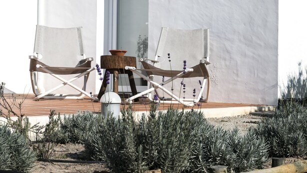 Un edificio blanco con terraza de madera, dos sillas blancas y una mesa pequeña con un cuenco y flores.