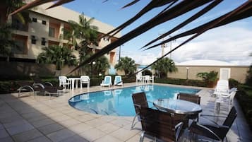 Una piscina con sillones y mesas, rodeada de palmeras y un edificio con balcones.