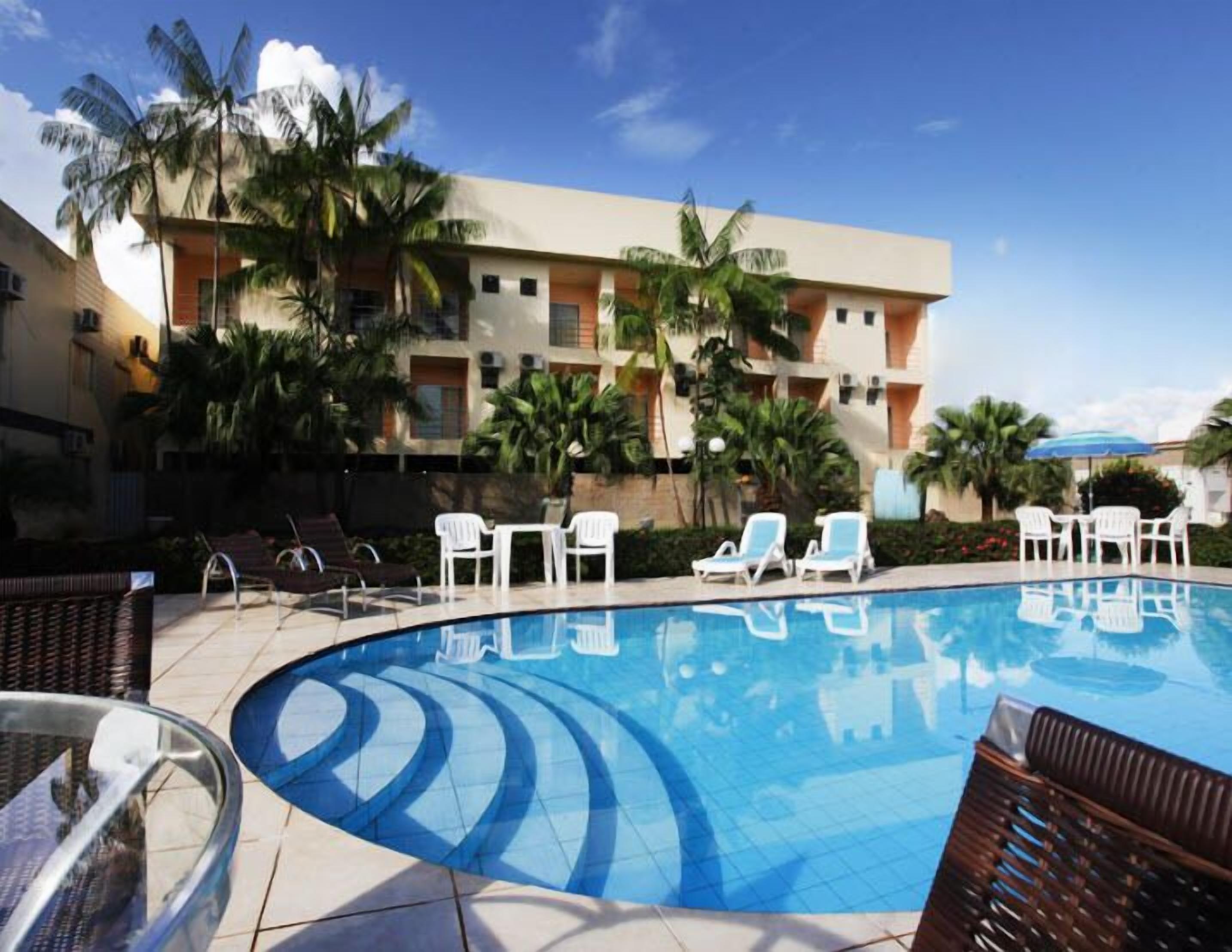 A swimming pool with lounge chairs and a building with balconies and palm trees in the background.