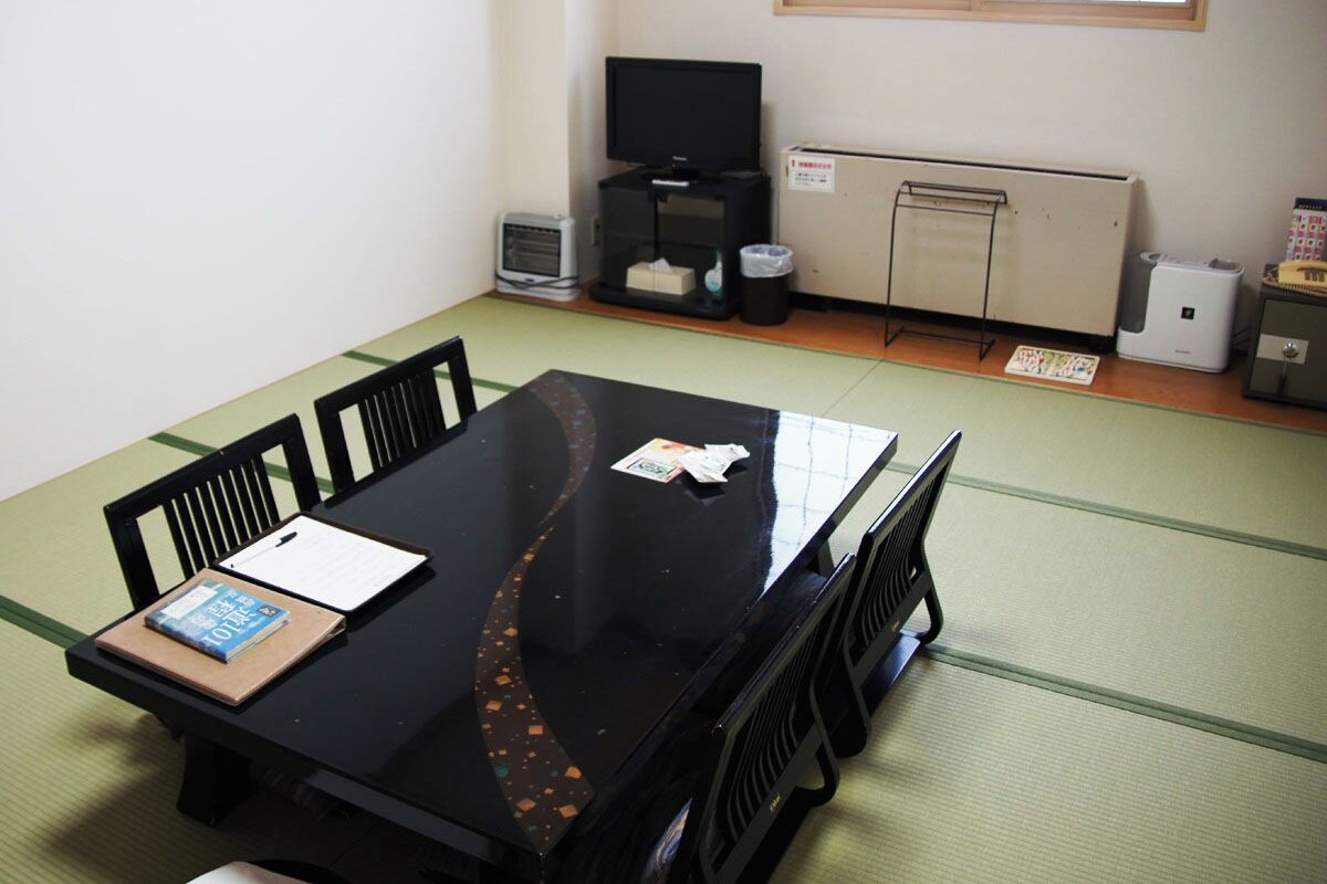 A room with tatami flooring, a black table with chairs, a television, a small heater, and a cabinet.