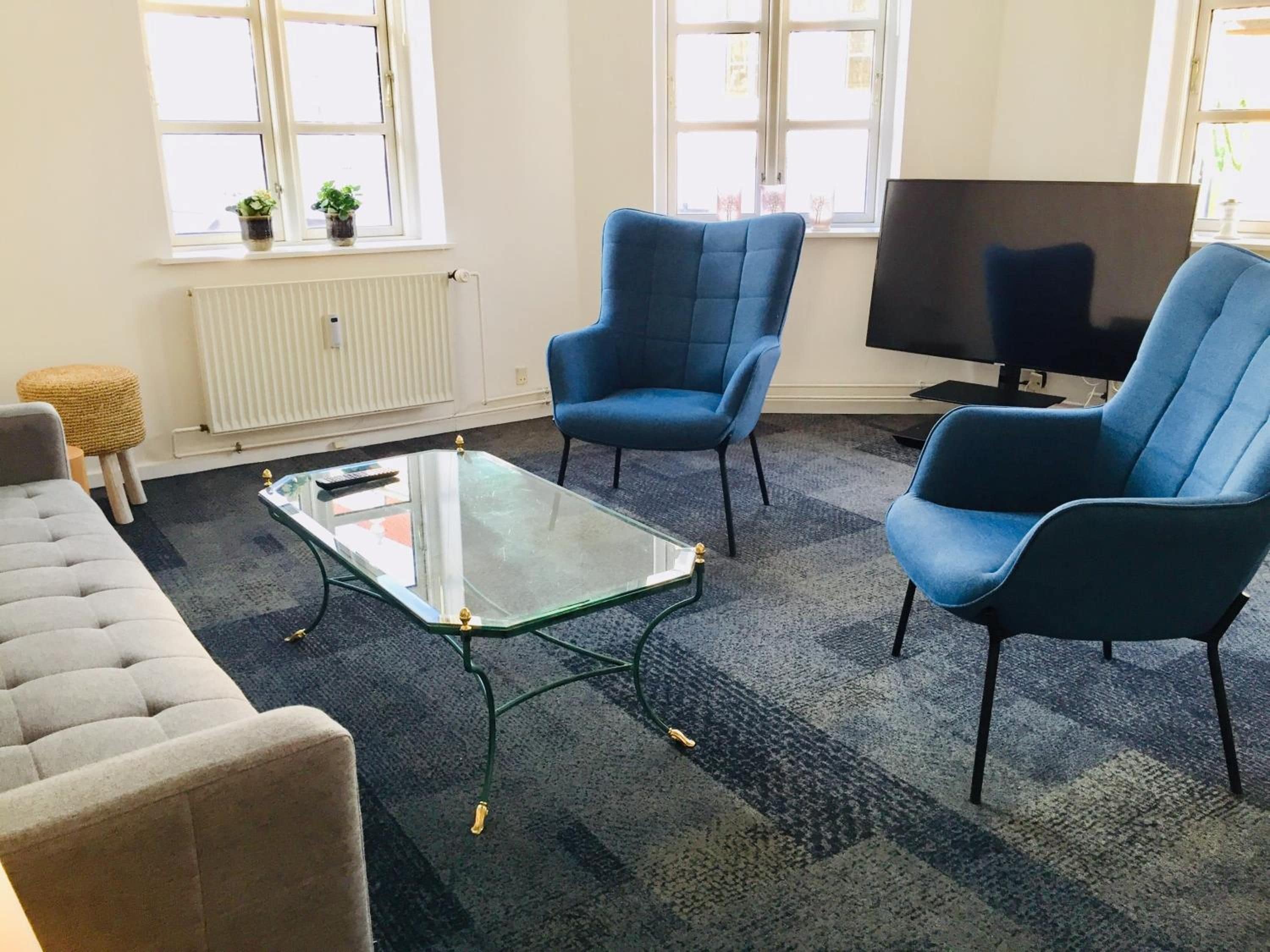 Ein Wohnzimmer mit einer Couch, zwei blauen Sesseln, einem Glastisch, einer Heizung, einem Fernseher und einem Fenster mit Pflanzen.
