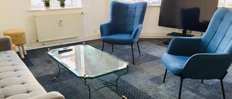 A living room with a sofa, two blue armchairs, a glass coffee table, a radiator, a TV, and a window with plants.