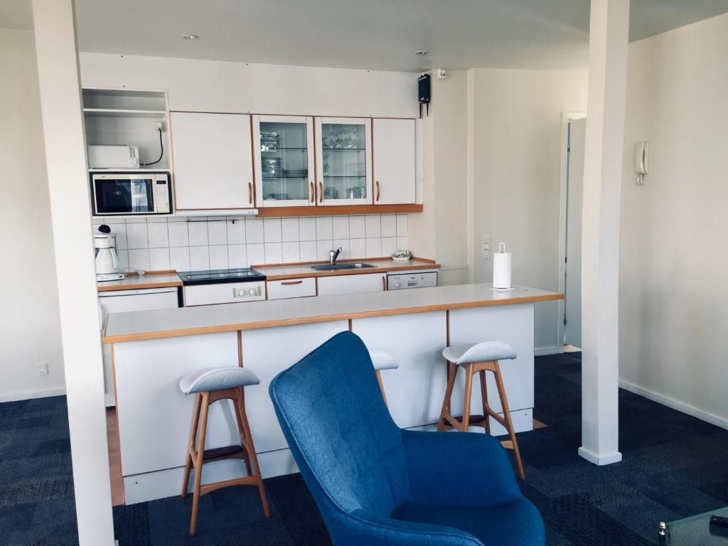 A compact kitchen with white cabinets, a microwave, and a coffee maker. There is a blue chair and two stools in the foreground.