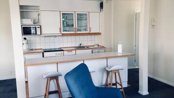 A compact kitchen with white cabinets, a microwave, and a coffee maker. There is a blue chair and two stools in the foreground.