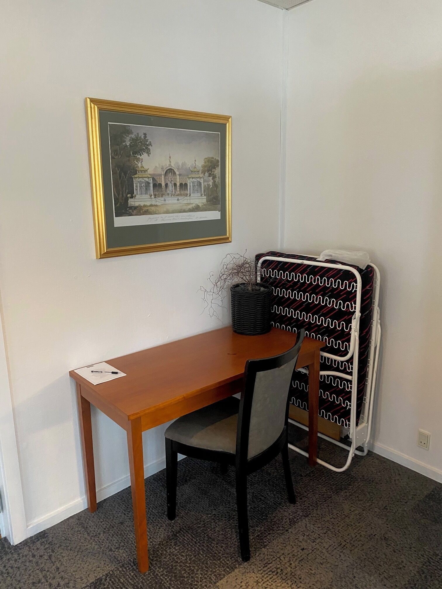Ett litet rum med ett skrivbord i trä, en stol, en inramad tavla på väggen och ett element.
