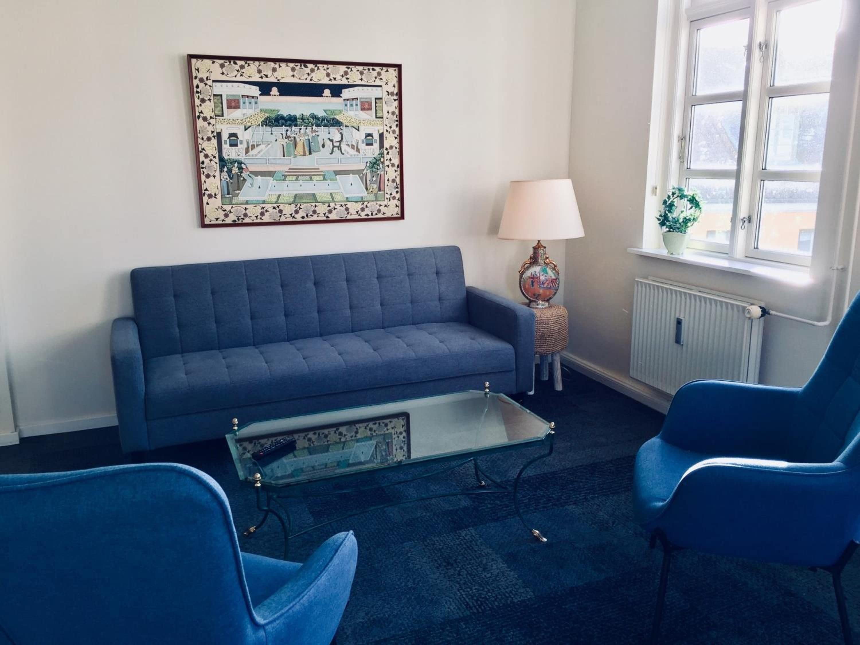 Une pièce avec un canapé bleu, deux fauteuils bleus, une table basse en verre, une lampe, un radiateur et une fenêtre donnant sur un beau paysage.