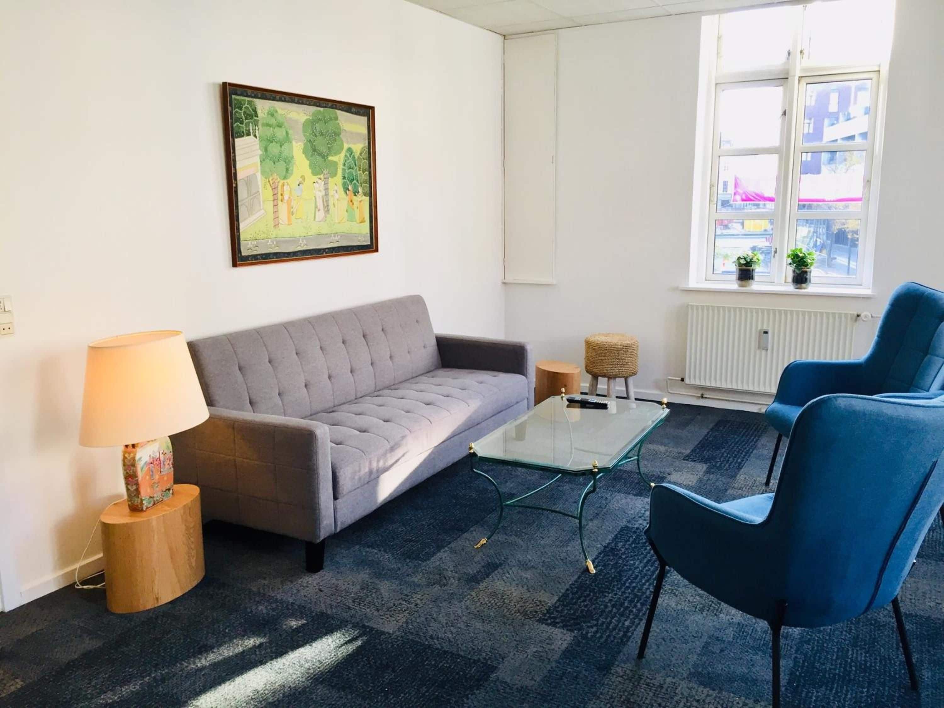 Una sala de estar con un sofá gris, un sillón azul, una mesa de centro de vidrio, una lámpara, un taburete redondo pequeño y un cuadro enmarcado en la pared.