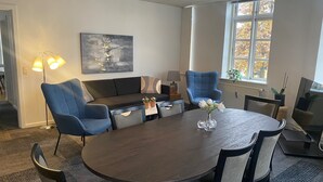 A modern dining area with a round table, blue chairs, a sofa, and a painting on the wall. - City Hotel Nattergalen Apartments (Odense)