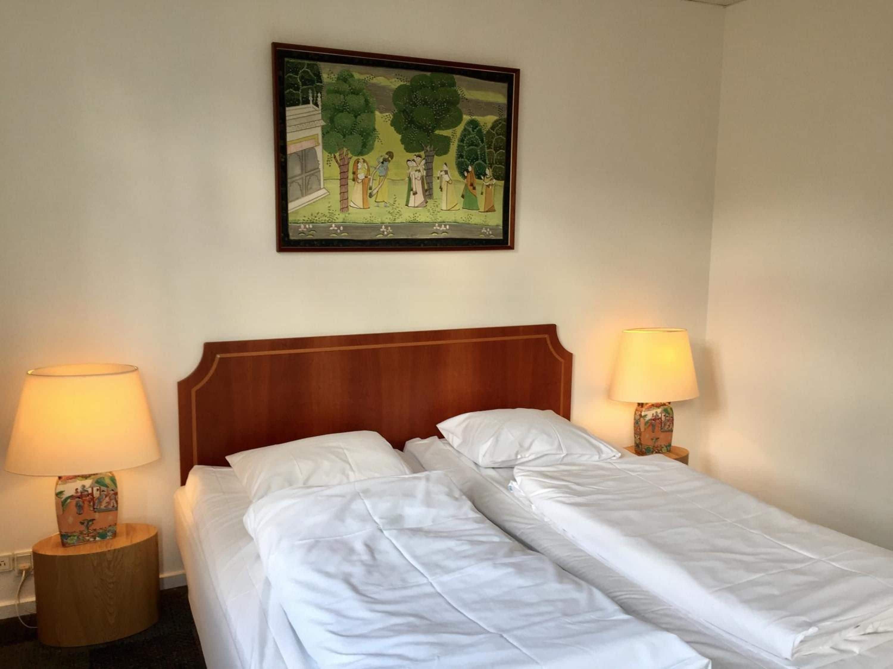 A hotel room with a double bed, two bedside lamps, a framed picture on the wall, and a small wooden side table.