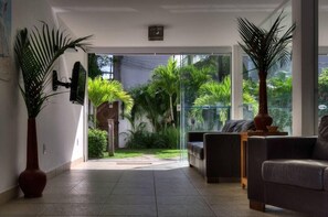 A modern living room with a glass door leading to a garden, a sofa, a coffee table, and a large potted plant. - Pousada Bahia 10 (Morro de Sao Paulo)
