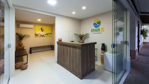 A reception area with a wooden reception desk, a wall-mounted logo, a potted plant, and a bench. - Pousada Bahia 10 (Morro de Sao Paulo)