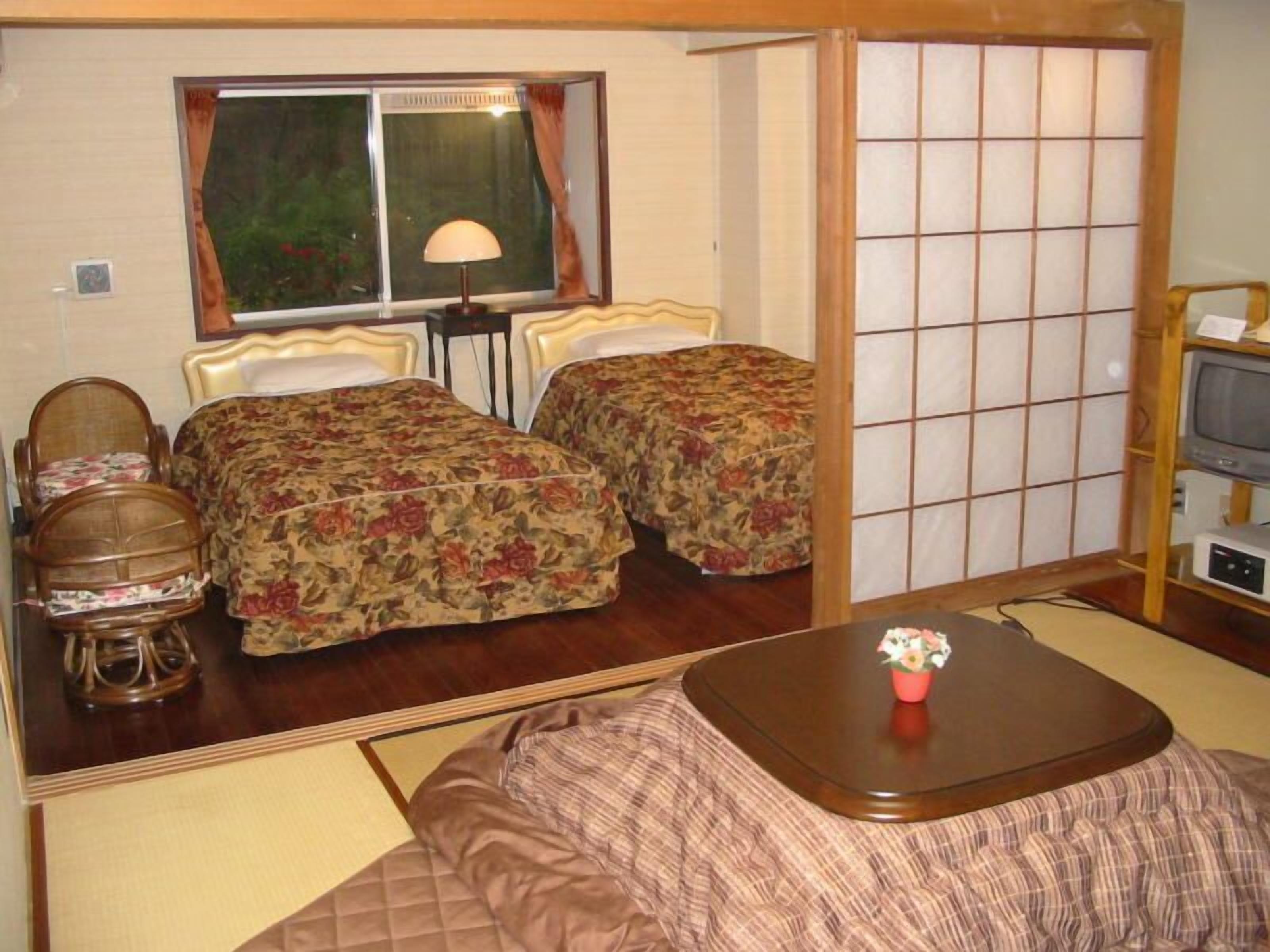 A room with two beds, a table, a chair, a TV, and a window with curtains.