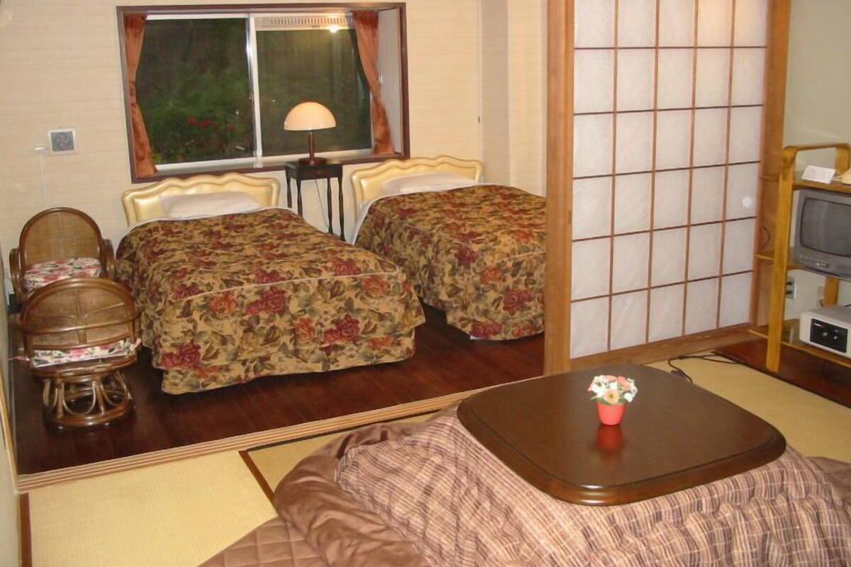 A room with two beds, a table, a chair, a TV, and a window with curtains.