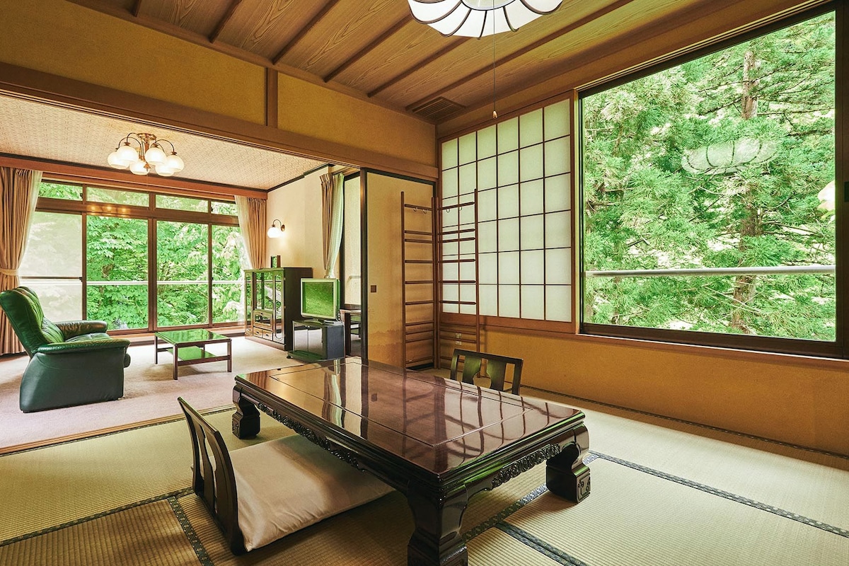A traditional Japanese room with tatami flooring, a low table, a green sofa, and a large window with a view of trees.