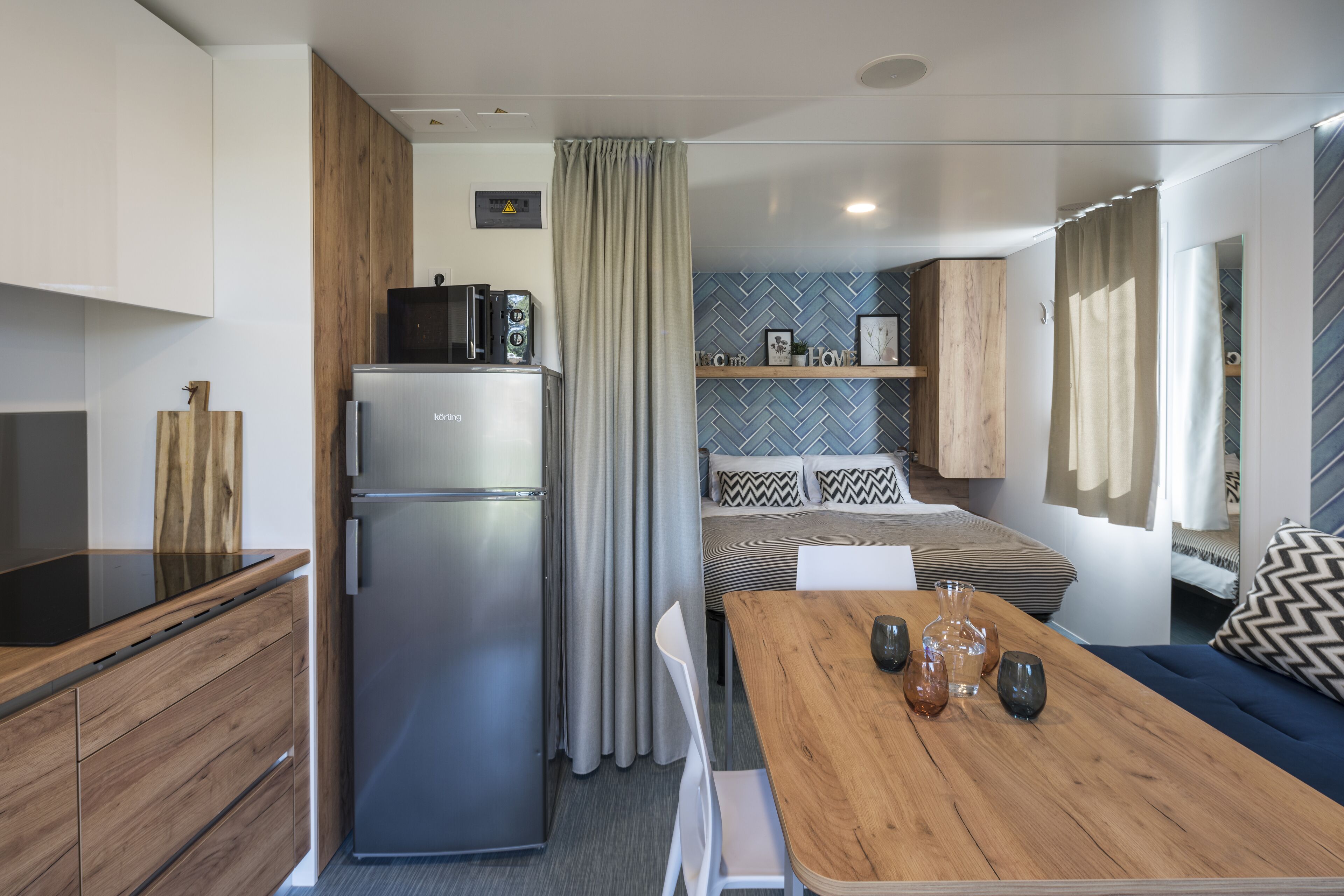 A compact kitchen with a refrigerator, microwave, and wooden cabinets. There is a dining area with a table and chairs, and a sofa in the background.