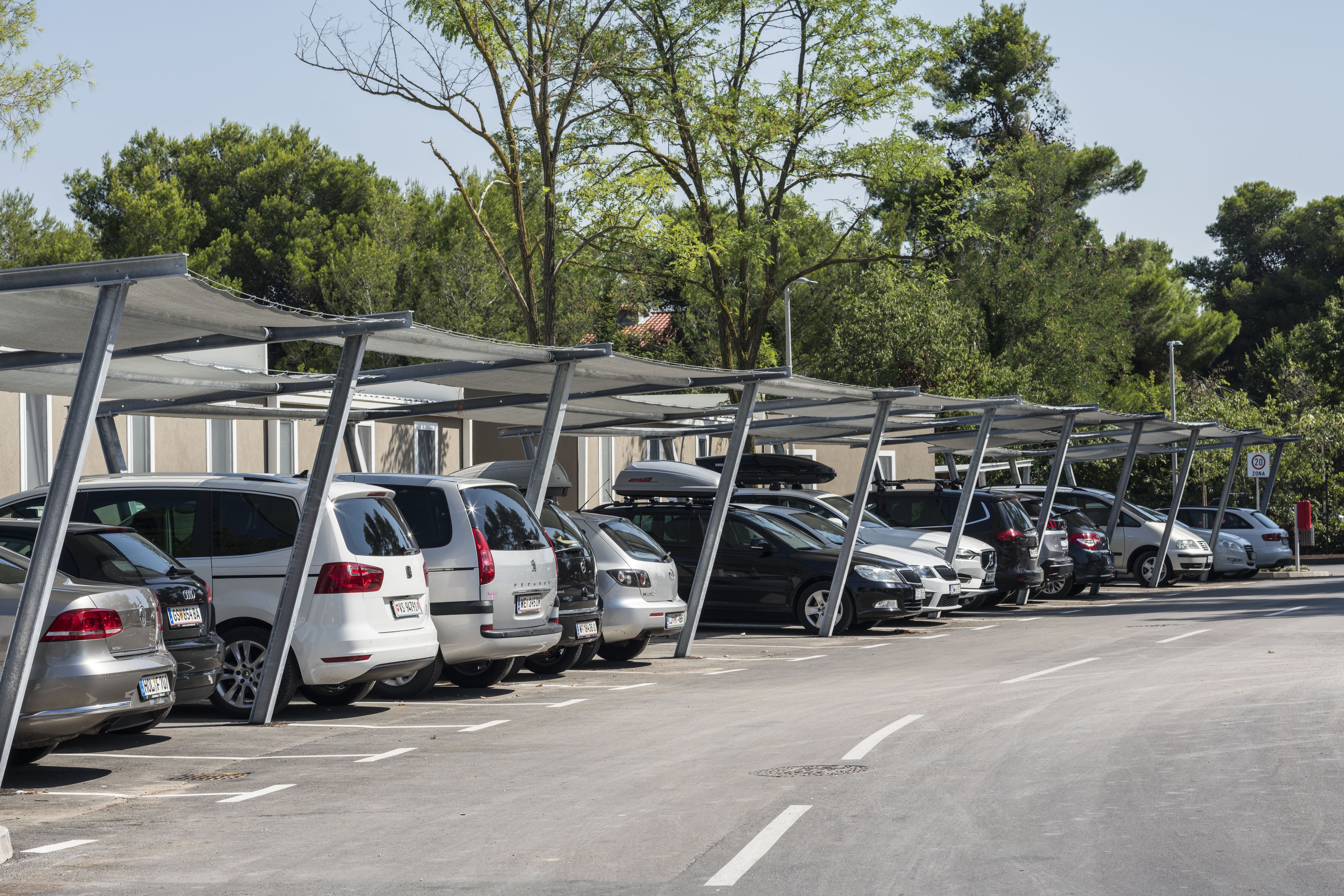 Una fila de coches aparcados bajo una zona cubierta para estacionamiento, con árboles al fondo.