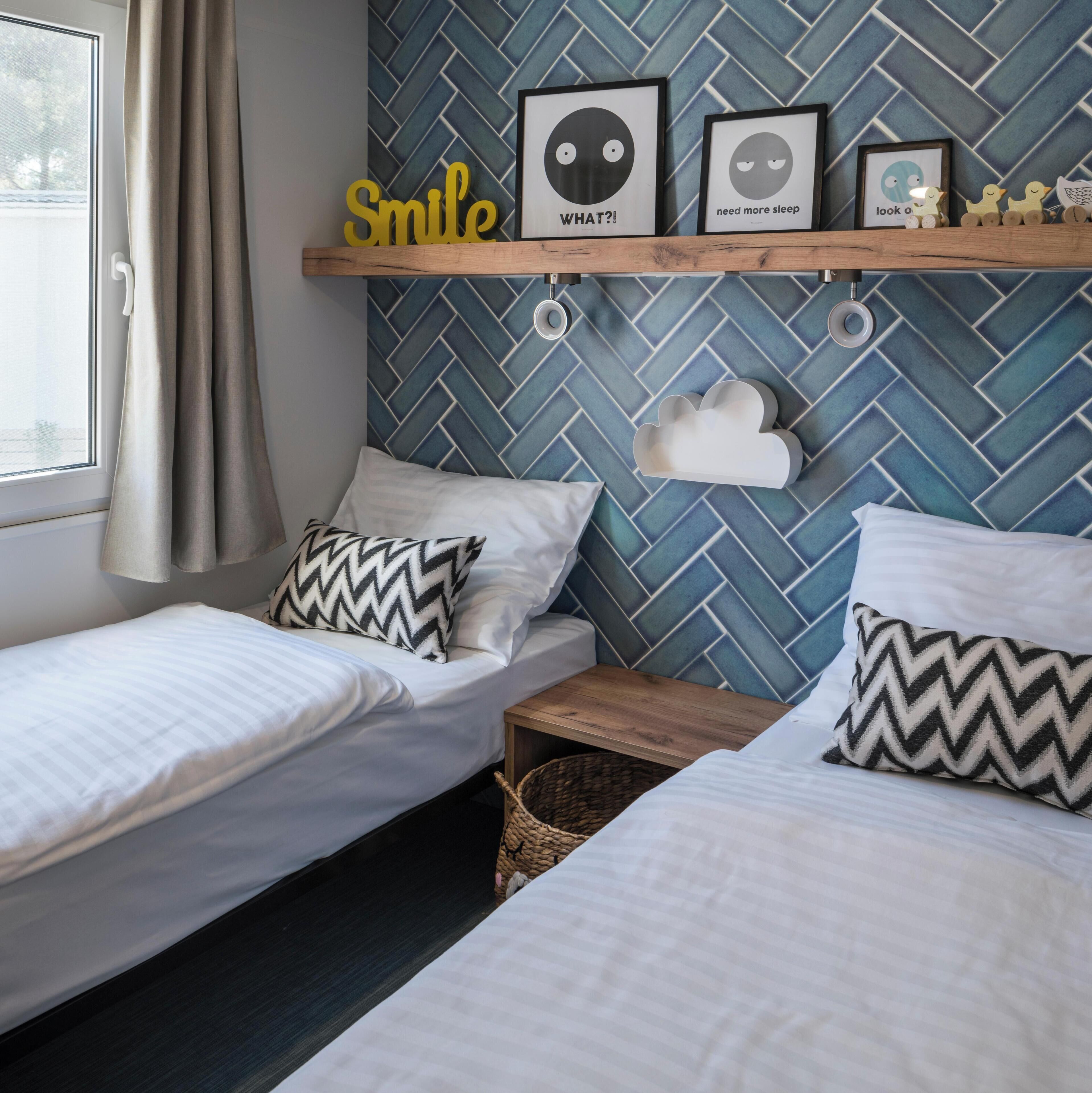 A bedroom with two beds, a window with curtains, a wall with a herringbone pattern, and a shelf with framed pictures and decorative items.
