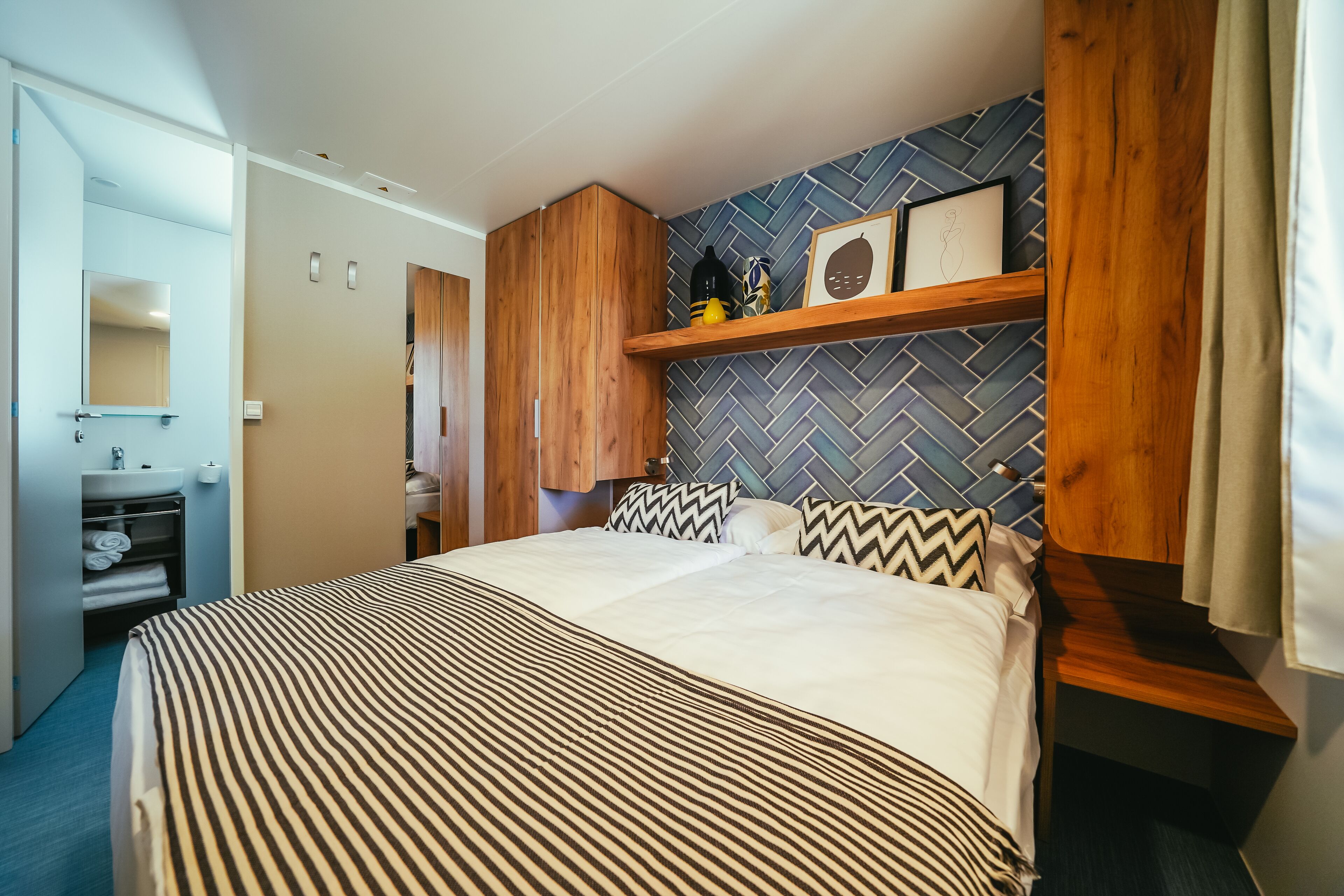 A hotel room with a large bed, wooden headboard, a small wooden shelf with decorative items, a mirror, and a bathroom with a sink and towel rack.