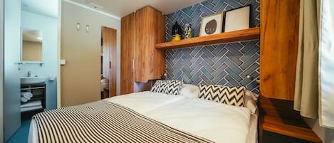 Ett hotellrum med en stor säng, ett träbeklätt sänggavel, en liten trähylla med dekorativa föremål, en spegel och ett badrum med handfat och handduksställ.